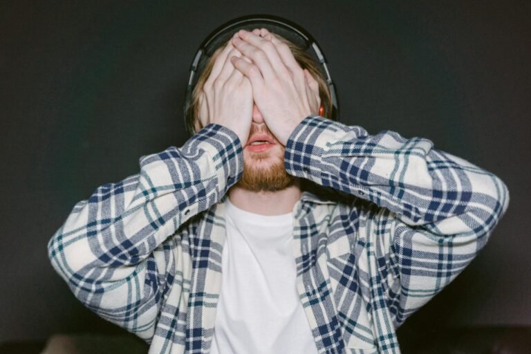 Young man wearing headphones and plaid shirt covering eyes, expressing stress or fatigue indoors.
