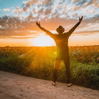 A man raises arms in freedom at sunrise in a rural field, expressing joy and connection with nature.